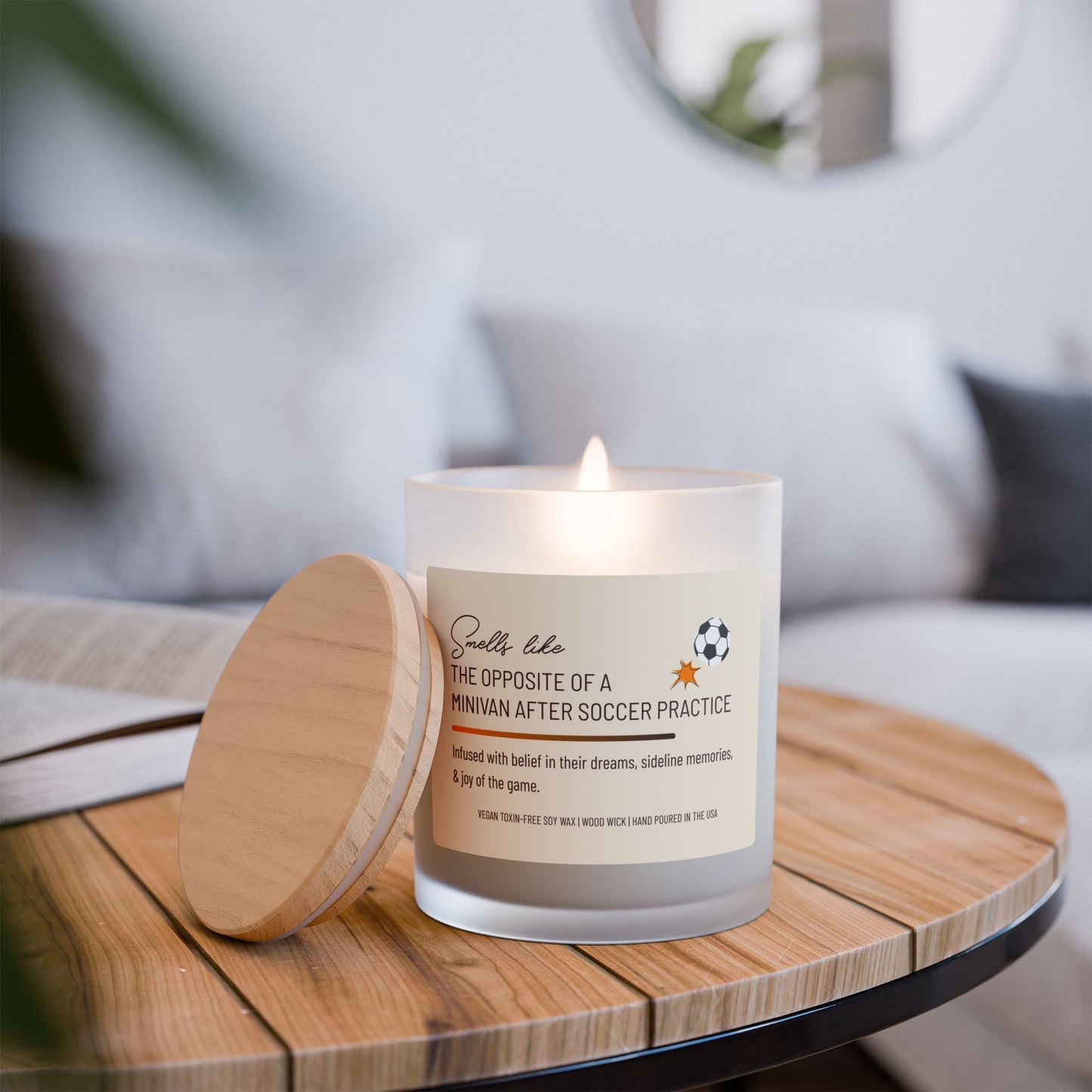 Frosted glass candle with wooden lid leaned against side of lit candle sitting on a wooden topped side table by a couch, labeled "Smells like the opposite of a minivan after soccer practice," featuring a small soccer ball graphic. This funny candle makes a great gift for women.