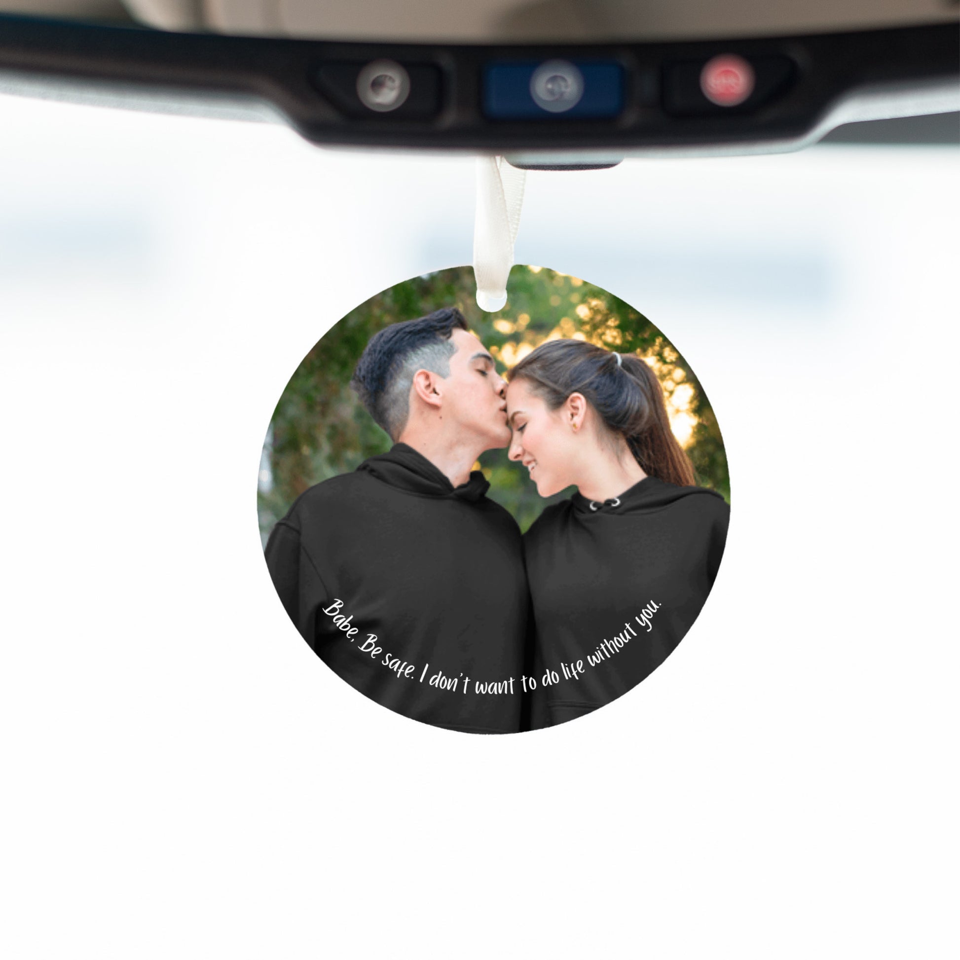 Car mirror hanging accessory ornament hanging below a mirror with a white background visible through the front windshield of the vehicle. Ornament features a personalized photo of a couple in black hoodies shares a tender moment outdoors as the man kisses the woman's forehead. Text below reads, "Babe, be safe, I don't want to do life without you." These make great gifts for women and men.