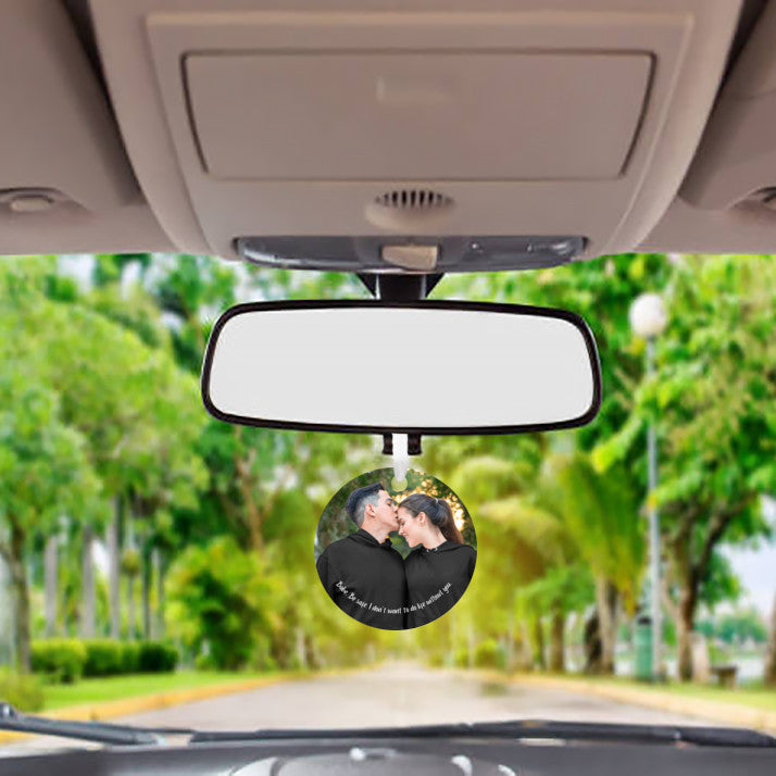 Car mirror hanging accessory ornament hanging below a mirror with the view of trees along the road through the front windshield of the vehicle in the background. Ornament features a personalized photo of a couple in black hoodies shares a tender moment outdoors as the man kisses the woman's forehead. Text below reads, "Babe, be safe, I don't want to do life without you." These make great gifts for women and men.