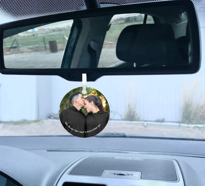 Car mirror hanging accessory ornament hanging below a mirror with the view of a white building in front of where the vehicle is parked seen through the front windshield of the vehicle in the background. Ornament features a personalized photo of a couple in black hoodies shares a tender moment outdoors as the man kisses the woman's forehead. Text below reads, "Babe, be safe, I don't want to do life without you." These make great gifts for women and men.