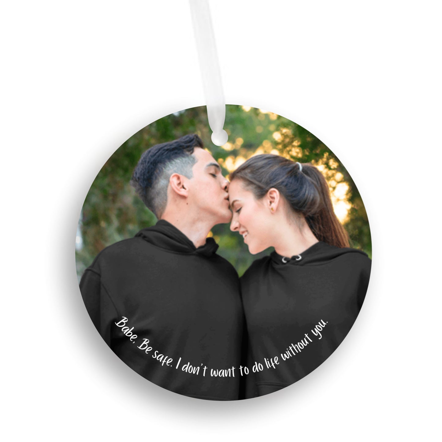 Car mirror hanging accessory ornament with white ribbon against a white background. Ornament features a personalized photo of a couple in black hoodies shares a tender moment outdoors as the man kisses the woman's forehead. Text below reads, "Babe, be safe, I don't want to do life without you." These make great gifts for women and men.