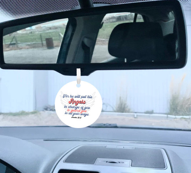 View from right side inside vehicle of a round white car mirror hanging accessory ornament with the Bible verse Psalm 91:11, featuring angel wings. The text, in blue and red, conveys protection and guardianship. Makes a great gift for women, men, teenagers, and anyone who drives a lot in their work.
